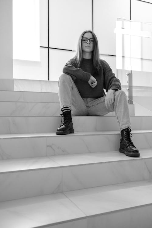 Young female in casual wear and eyeglasses sitting on stairs in house while looking at camera