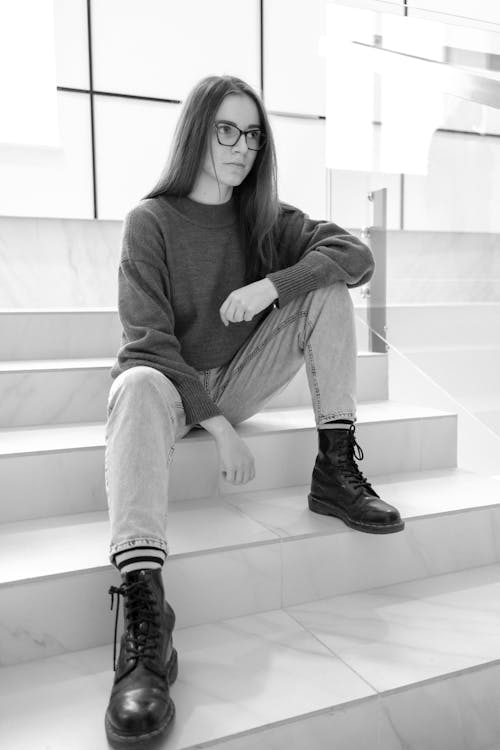 Black and white of young female in leather boots and jeans sitting on staircase while looking forward in building