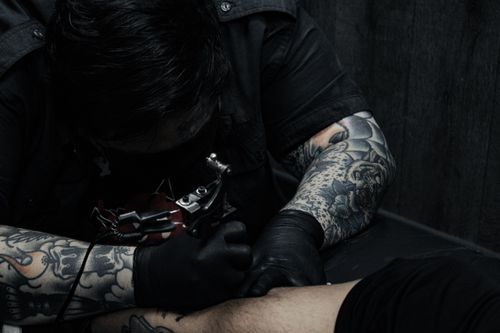 Free Tattoo artist focused on creating an intricate arm tattoo with a tattoo machine in a dimly lit studio. Stock Photo