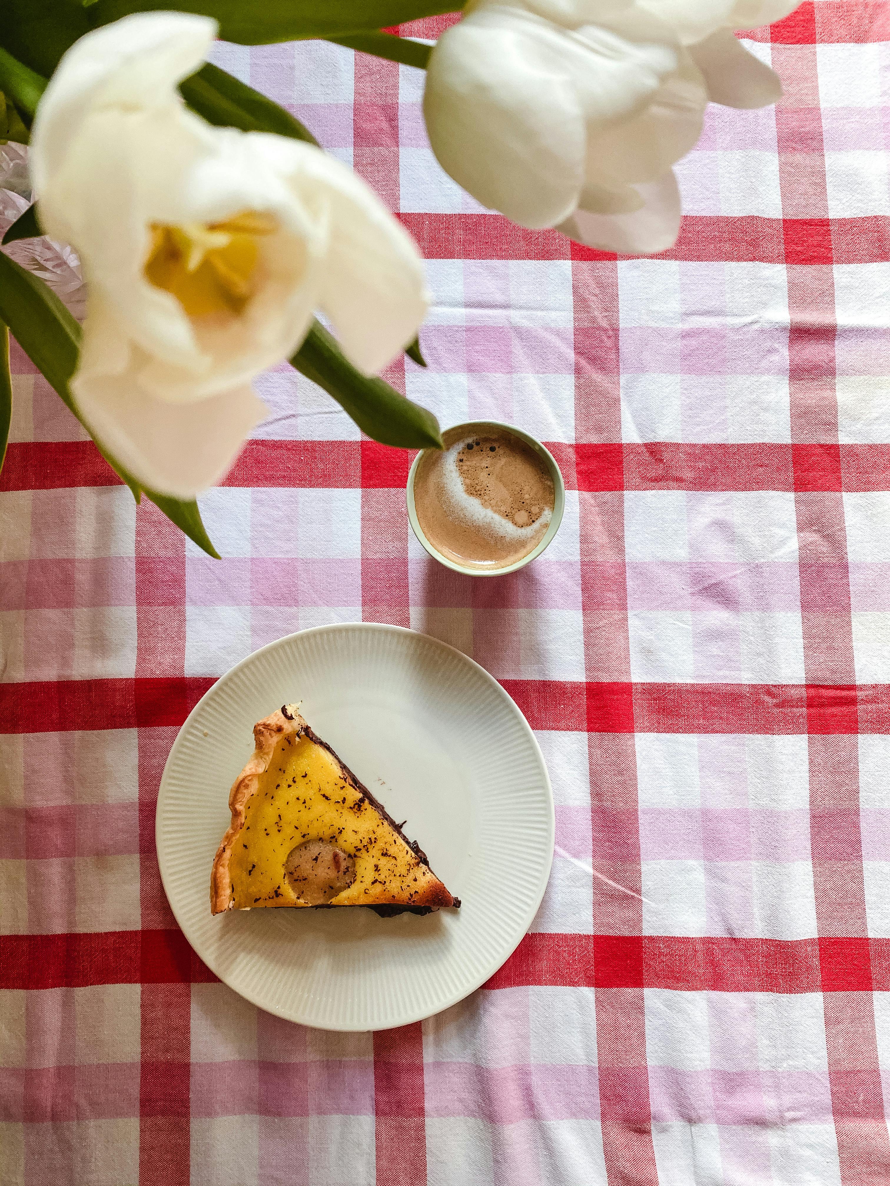 Slice of pie with coffee on a checkered tablecloth beside white flowers. Ideal breakfast setting.