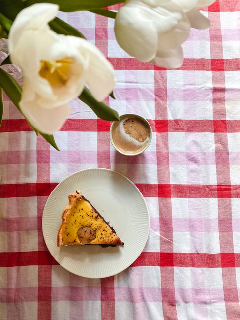 Slice of pie with coffee on a checkered tablecloth beside white flowers. Ideal breakfast setting.