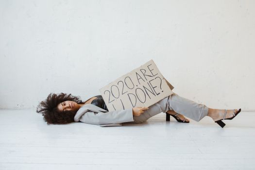 A woman lying on the floor holding a sign reading '2020, Are We Done?' expressing fatigue.