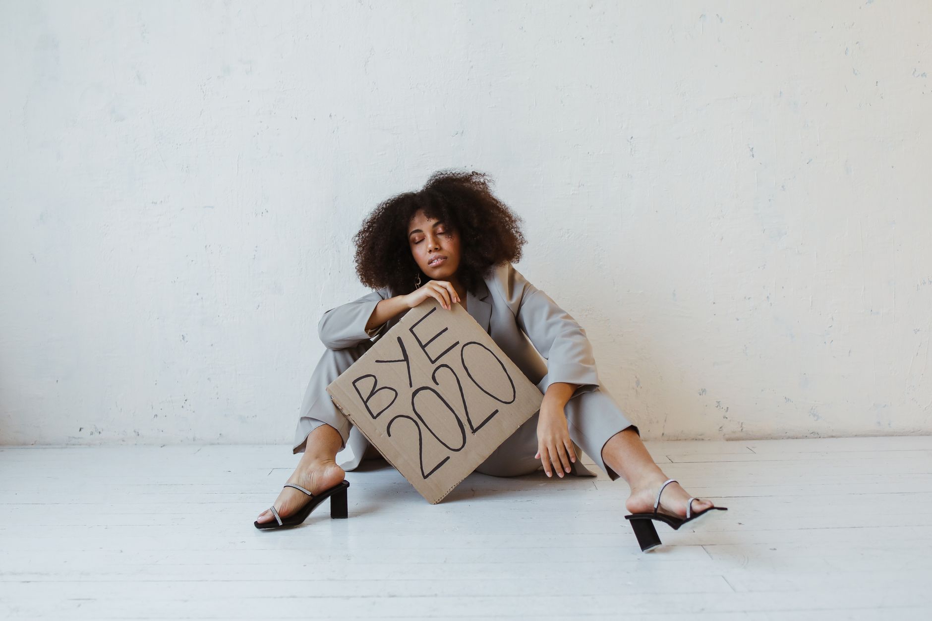 Woman sitting on floor holding 'Bye 2020' sign, expressing exhaustion and relief.