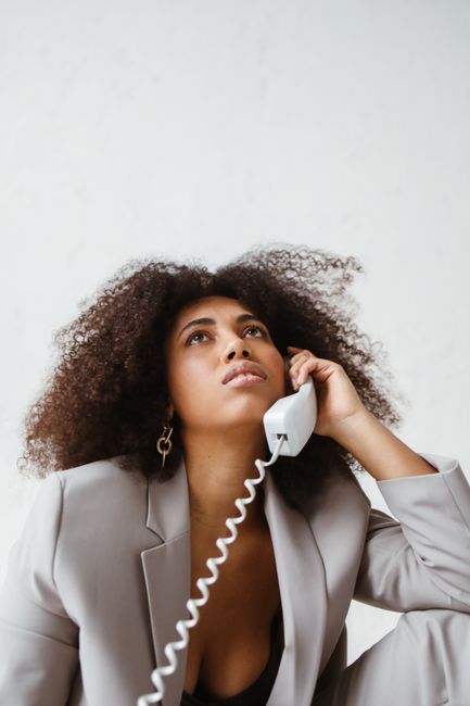 Stylish woman with curly hair in a beige suit talking on a vintage corded phone indoors.