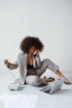 Smiling woman in a stylish suit sits on the floor multitasking with a phone and laptop.