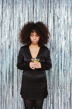 Portrait of a woman in black holding a drink against shiny tinsel backdrop.