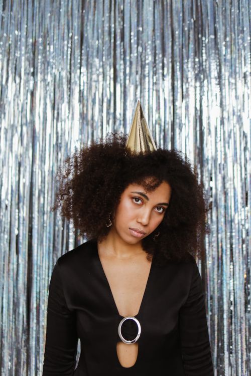 Free A young woman with afro hair in a black outfit and party hat stands against a sparkling backdrop. Stock Photo