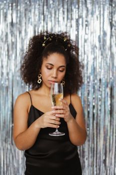 A thoughtful woman in a black dress holds a champagne glass, set against a shimmering background.