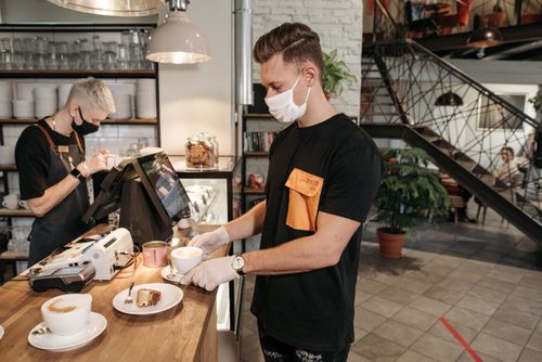 Free Two baristas in masks serving coffee in a chic, indoor café setting. Stock Photo