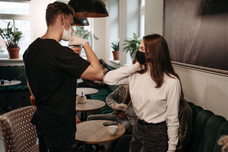 Two friends greeting with elbow bump in a café, wearing masks for safety.