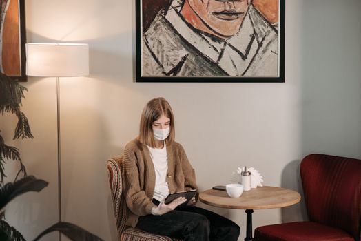 A woman wearing a face mask sits indoors using a tablet, emphasizing modern lifestyle and safety.