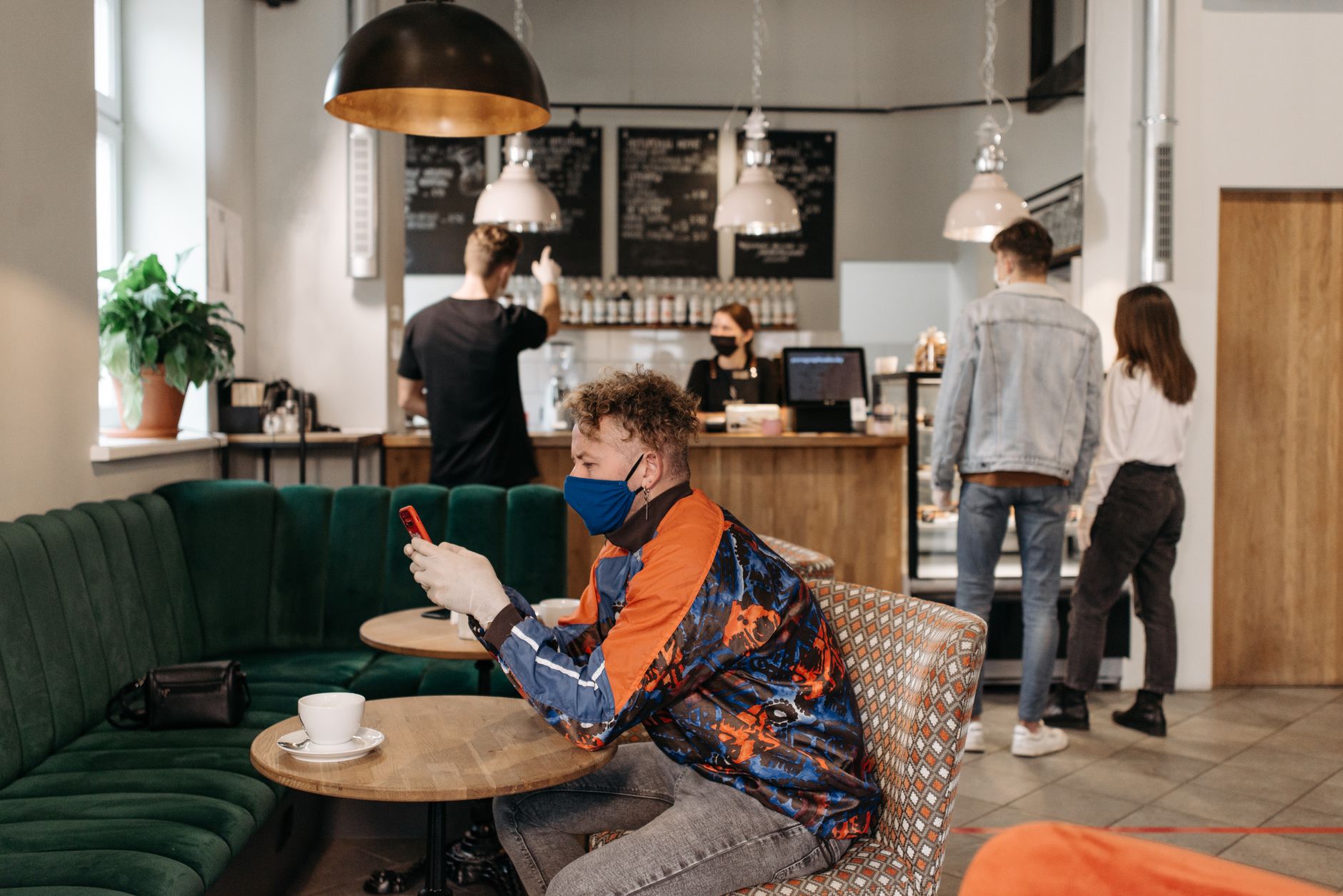 A man wearing a face mask uses his phone in a stylish café with people around.