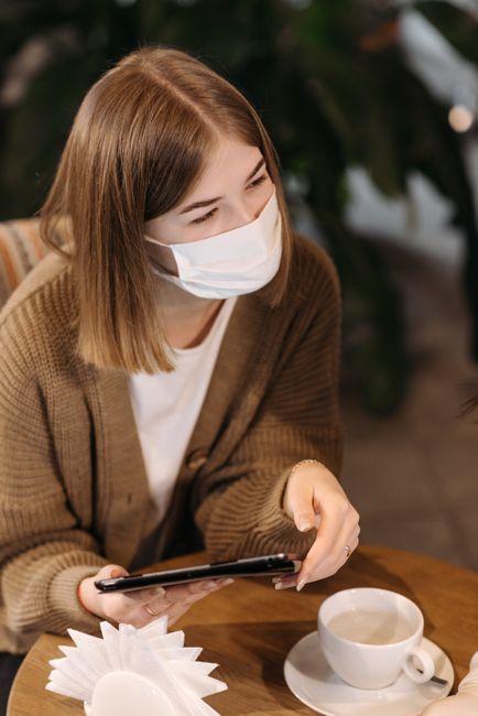 Woman wearing a face mask and using her smartphone in a cozy coffee shop.