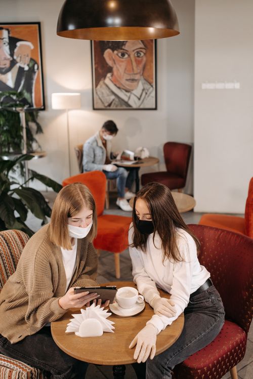 Free Two women in face masks enjoy coffee inside a cozy cafe setting, embodying a pandemic-era lifestyle moment. Stock Photo