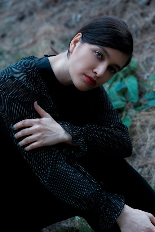 Unemotional attractive female in black blouse sitting on ground in nature and looking at camera