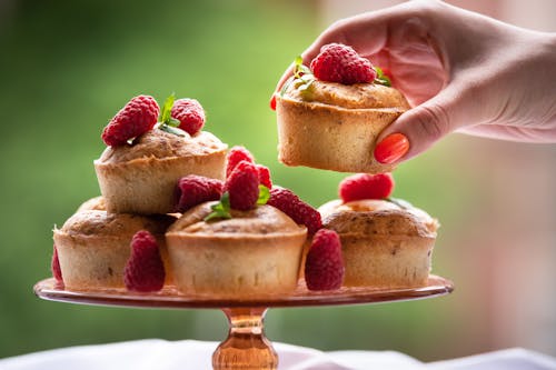 Crop faceless lady taking delicious homemade cakes placed on table with white tablecloth and decorated with raspberries