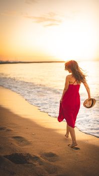 Back view of faceless female in red dress holding straw hat while strolling on seashore under bright sunlight