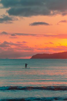 A person paddleboarding at sunset, creating a tranquil silhouette against a vibrant sky and calm ocean.