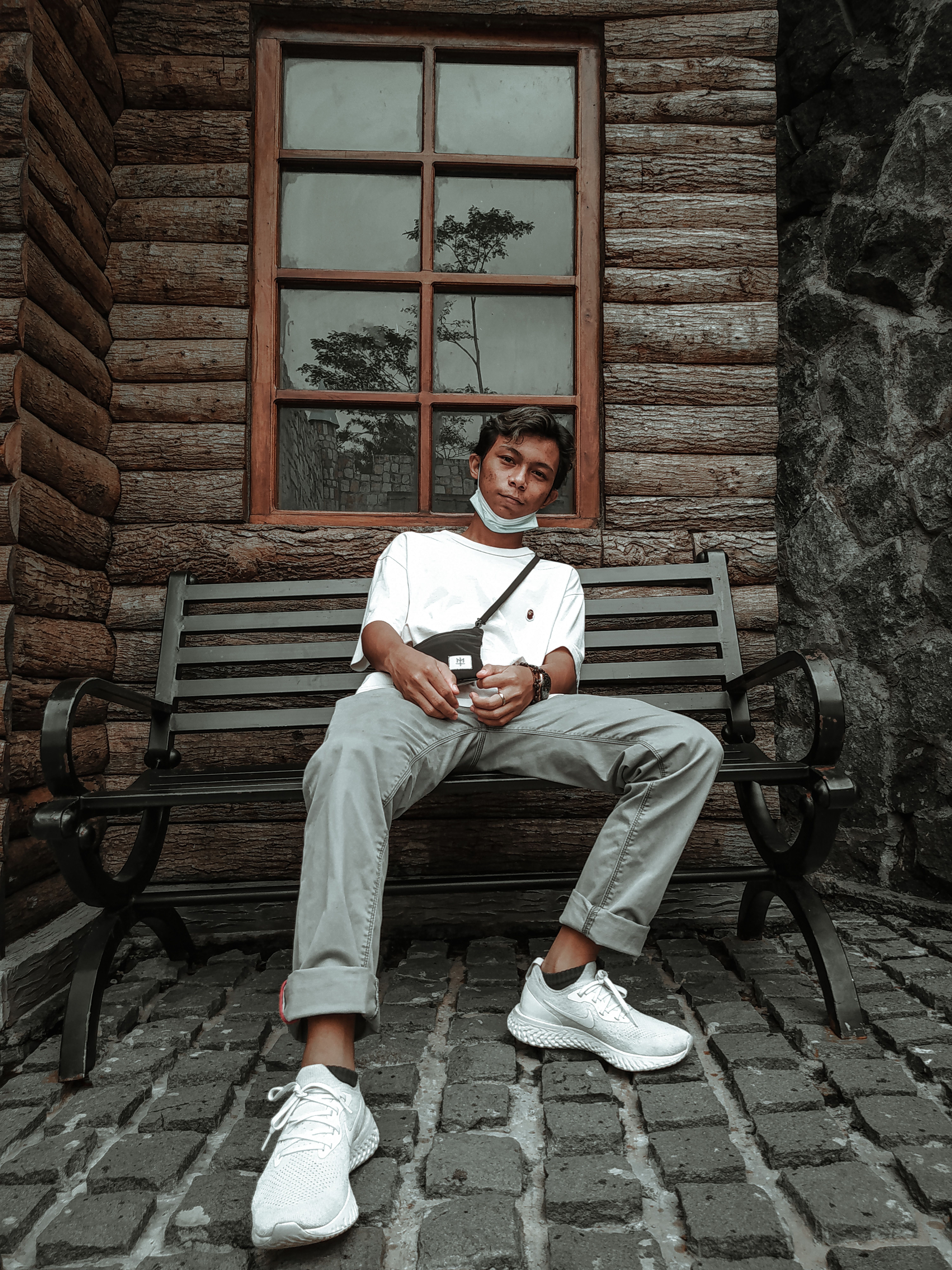 Full body of self assured young Asian male millennial in trendy outfit and sneakers chilling on bench near wooden house in town and looking at camera