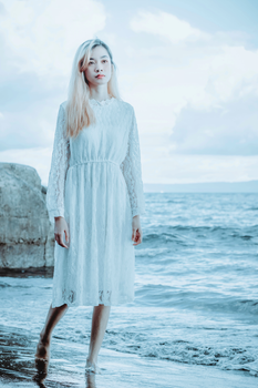 Full body thoughtful barefoot young Asian female in white dress walking along wet sandy beach near waving sea water and looking away