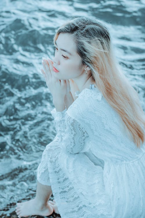 High angle of calm pensive young Asian female in white dress sitting on wet seashore and looking away