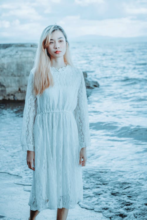 Young attractive Asian female in long white dress with long blond hair standing on seashore and looking away