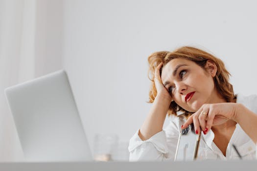 A woman with short hair and red lips looks thoughtful holding a smartphone beside a laptop.