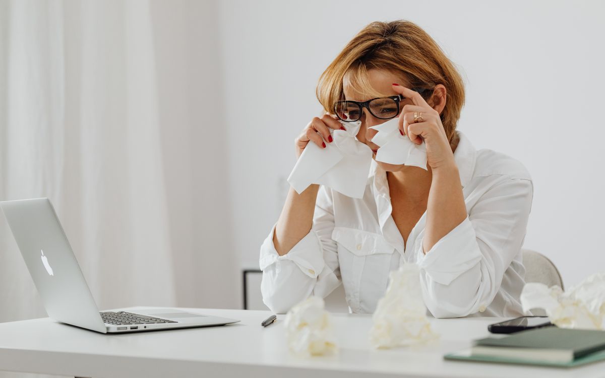 A woman in glasses is wiping tears while seated at a desk with a laptop and tissues.