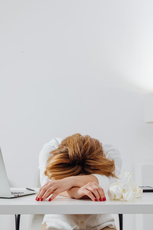 Free Picture of a tired woman with red nails resting her head on a white desk in a bright office setting. Stock Photo- Blog: Simple Ways to Feel Better and Stop Burnout Before It Starts