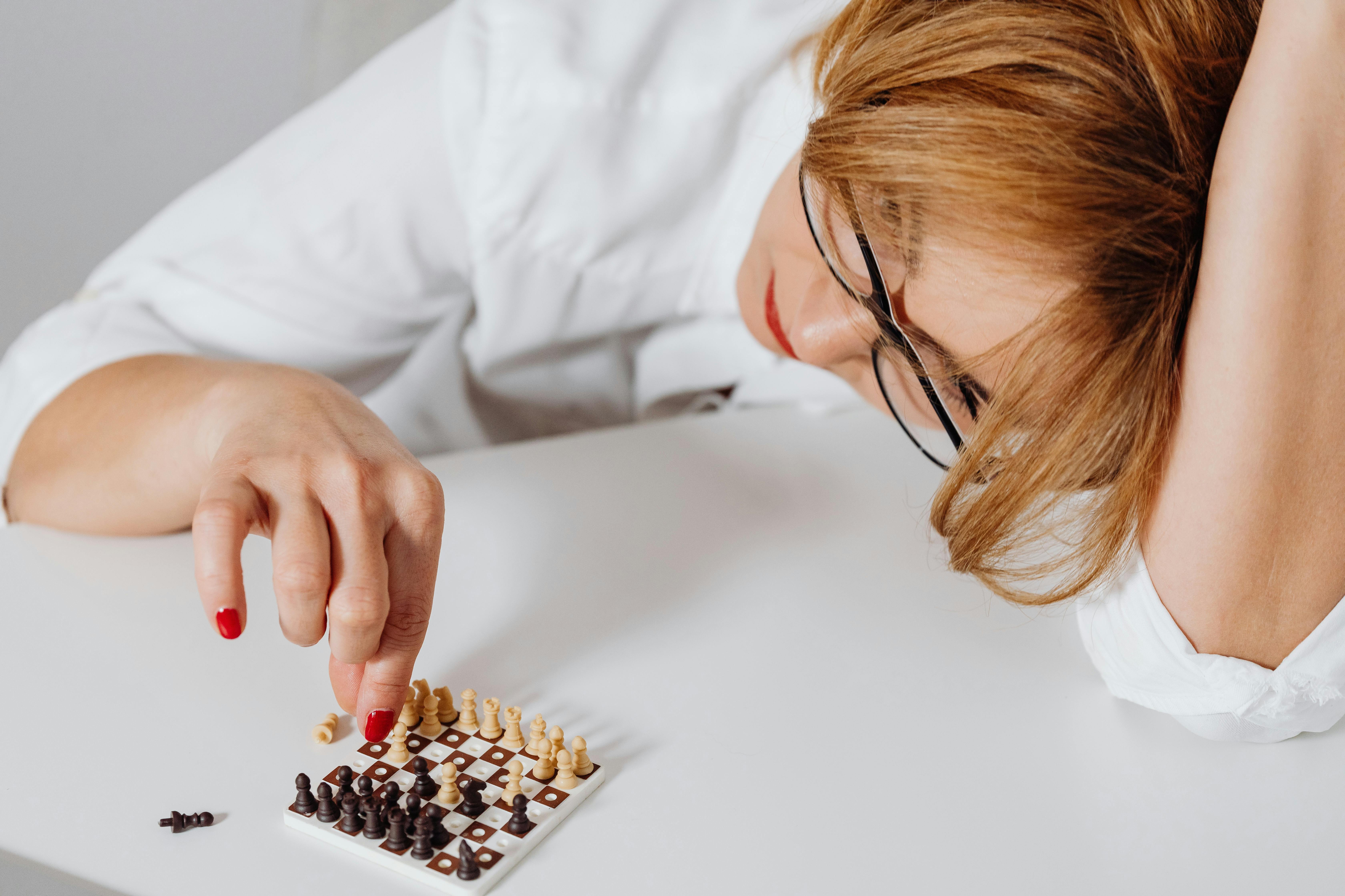Close-up of a woman in glasses focusing on a miniature chess game indoors.