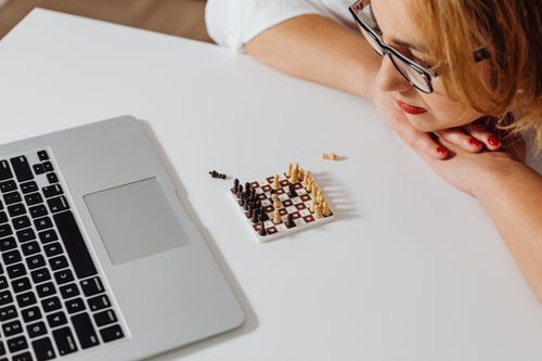 Free Focused woman looking at miniature chess set on white table with laptop nearby. Stock Photo