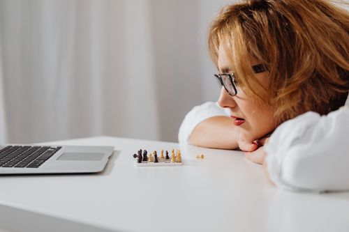 Free A woman thoughtfully observes a miniature chess set next to a laptop, focusing on the game. Stock Photo
