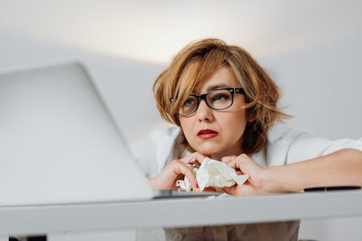 A woman looking stressed, sitting at a desk with a laptop, crumpled papers, and eyeglasses.
