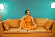 Full body of focused barefoot female wearing casual clothes sitting with bent leg and pot with flower near face on sofa near green wall in room