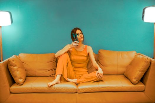 Full body of focused barefoot female wearing casual clothes sitting with bent leg and pot with flower near face on sofa near green wall in room