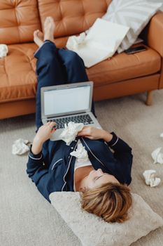 A woman in pajamas working remotely on a laptop at home, surrounded by crumpled papers.