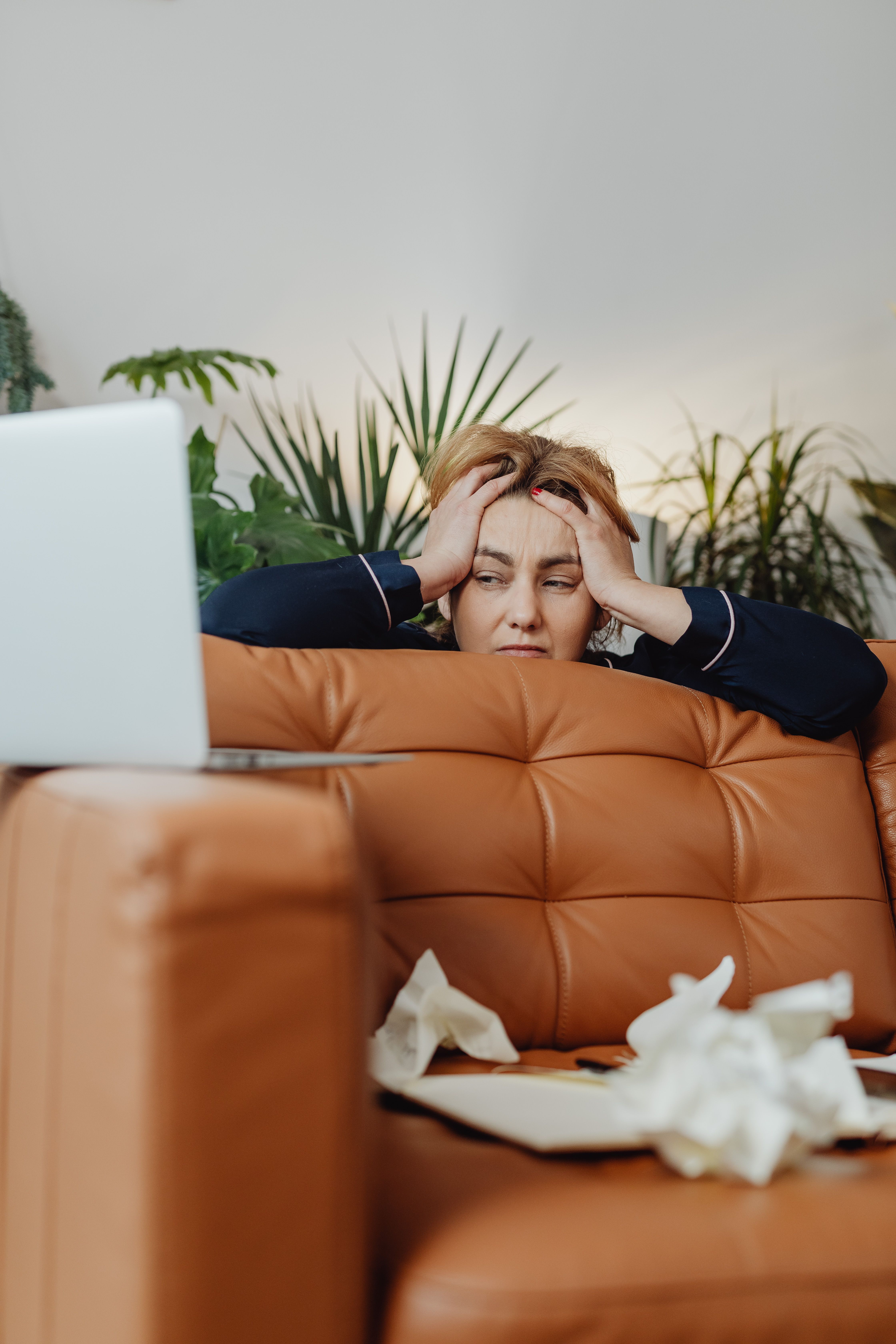 Frustrated woman holding her head with a laptop in front and plants surrounding her, depicting stress.