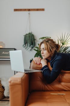 A woman lies on a brown leather couch using a laptop, surrounded by plants indoors.
