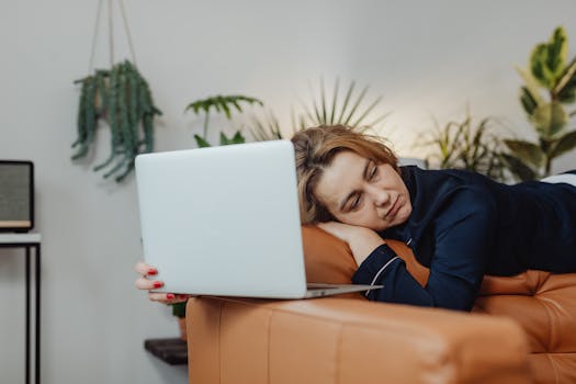 Woman sleeping on couch with laptop, surrounded by plants, indoors.