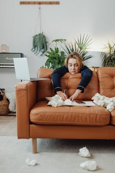 Woman lying over sofa, writing with crumpled papers around, showing stress and frustration.