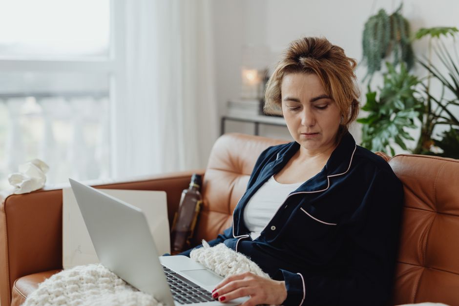 A woman sitting on a couch using a laptop, appearing stressed and overwhelmed.