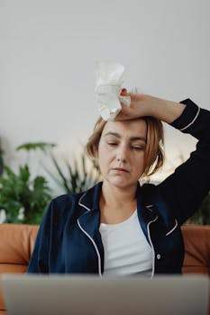 A tired woman holding crumpled paper sits indoors with plants and a laptop visible.