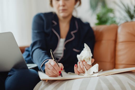 A woman writing in a notebook with tissues nearby, suggesting remote work or illness at home.
