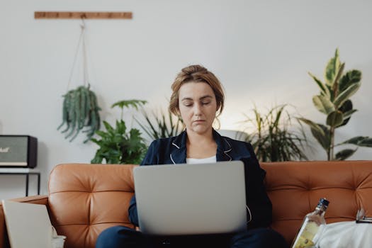 Woman in pajamas working on a laptop in a cozy living room surrounded by plants.