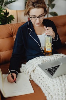 Woman sitting on couch writing in notebook, using laptop, holding gin bottle