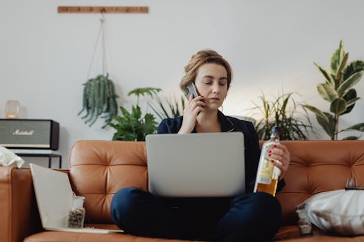 Woman multitasking at home with a laptop and phone, holding a bottle. Indoor setting with plants.