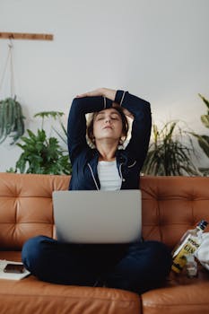 A woman in pajamas takes a stretch break while working from home on her laptop.