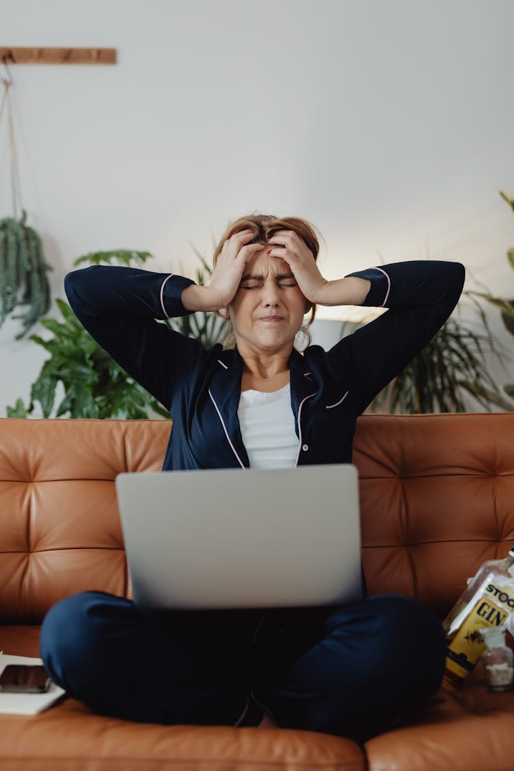 A Woman With A Headache Sitting On A Couch With Her Laptop