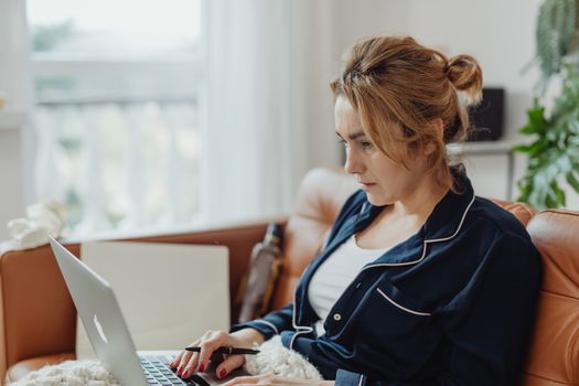 Woman in pajamas working on a laptop from a cozy home environment, focused on her task.