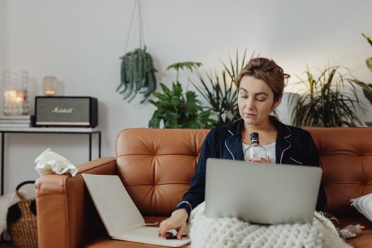 A woman in pajamas holding a bottle, using a laptop on a sofa in a cozy living room setting.